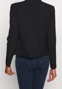 Černý zkrácený blazer se strukturovanými rameny a dlouhými rukávy, zkombinovaný s tmavě modrými džíny s klasickým pěti kapsovým designem.