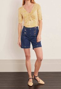 Žlutá blůza s vzorem a nabíranými rukávy, modré džíny s vysokým pasem s knoflíkovými detaily a béžové nazouvací boty.