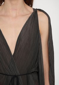 Robe enveloppante noire et texturée avec un décolleté en V profond, une taille à nouer et des plis verticaux distincts. Une manche est ouverte, révélant la peau en dessous.