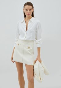 Camicia bianca con bottoni dorati, abbinata a una mini gonna bianca texture con tasche frontali e orlo sfrangiato, completata da un giubbotto.