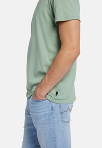 Maglietta in cotone verde chiaro con maniche corte, indossata con jeans in denim blu chiaro. Dettaglio di cucitura sottile sulla tasca dei jeans.