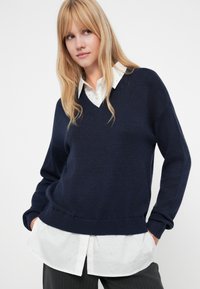 Navy v-neck sweater with a relaxed fit, ribbed cuffs and hem, layered over a white collared shirt with subtle polka dots.