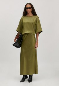 Olijfgroen satijnen tweedelige outfit met korte mouwen en een rok tot de knie. Gecompleteerd met een zwarte tas en enkellaarsjes.