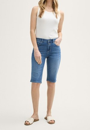 Kvinde iført en hvid ærmeløs top, blå knælange denimshorts og beige sandaler, der står mod en ensfarvet baggrund.