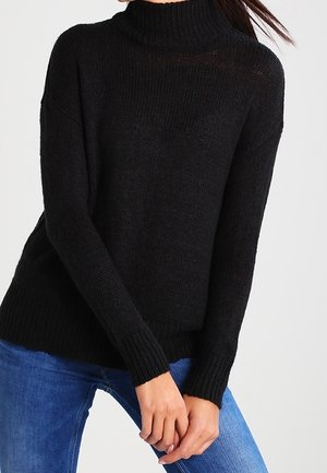 Mustat neulotut neuleet, joissa on korkea kaulus, ribbikädentyöt ja rento istuvuus. Yhdistetty sinisiin farkkuihin, korostaen rakennetta ja rentoa muotoilua.