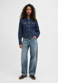 Tamsiai mėlynas džinsinis švarkas su dviem krūtinės kišenėmis, mygtukų uždarymu ir apykakle. Derinamas su plačiomis šviesiai mėlynomis kelnėmis ir juodais batais.