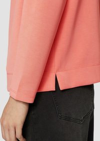 Koraliowa koszula z długim rękawem o prostym kroju, z wycięciem po boku i wykończeniem szwem. Noszona z dopasowanymi czarnymi dżinsami.