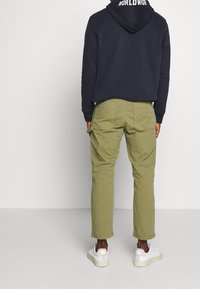 Persoon gekleed in olijfgroene broek, witte sneakers en een marineblauwe hoodie met de tekst "WORLDWIDE" op de capuchon, staand tegen een eenvoudige witte achtergrond.