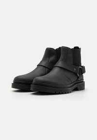 Botas negras de tobillo hechas de cuero suave con suela de goma, que presentan una correa ajustable y paneles laterales elásticos para facilitar su uso.