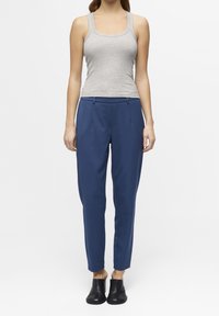 Een vrouw staat in een grijze tanktop en blauwe broek, gecombineerd met zwarte instapschoenen, tegen een eenvoudige witte achtergrond.