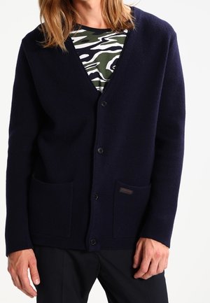 Persoon draagt een marineblauwe, met knopen sluitende cardigan met twee voorzakken over een groen, wit en zwart patroon top, gecombineerd met een zwarte broek.