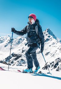 Person, die in schwarzer, isolierter Kleidung und blauen Skischuhen mit einem Skistock auf schneebedecktem Gelände fährt, im Hintergrund Berge.