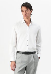 Man wearing a white button-up shirt with dark buttons, light gray pleated trousers, and a black leather belt, standing with one hand in pocket.