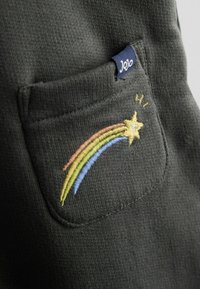 Tamsiai pilka medžiaga su kišenę, kuri puošia daugiakampė siuvinėjimo vaivorykštė ir žvaigždžių dizainas. Etiketėje kampe užrašyta "Jojo".