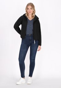 Juodas zip-up džemperis su aukšta apykakle, derinamas su tamsiai mėlynu dryžuotu marškinėliu ir siaurais tamsiais džinsais. Modelis vilki baltus sportinius batelius.