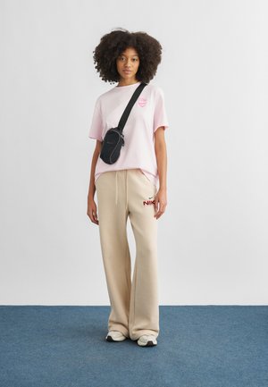 Kobieta o kręconych włosach w różowym t-shircie, beżowych spodniach dresowych Nike, białych trampkach i czarnej torebce na ramię, stojąca na niebieskiej podłodze.