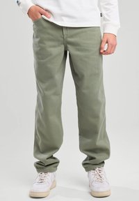 Pantalones de pierna ancha en verde oliva, confeccionados en una tela suave y texturizada. Presentan un ajuste clásico, bolsillos laterales y puños elásticos. Llevados con zapatillas blancas.