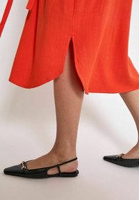 Scarpe slingback nere con punta affilata e suola piatta, con un dettaglio dorato sul davanti. Indossate con una gonna arancione che presenta uno spacco laterale.