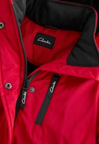 Rød isoleret jakke med en sort krave, der har en lynlås-lomme og fremtrædende Clarks-branding på mærkerne. Glat, vandafvisende materiale.