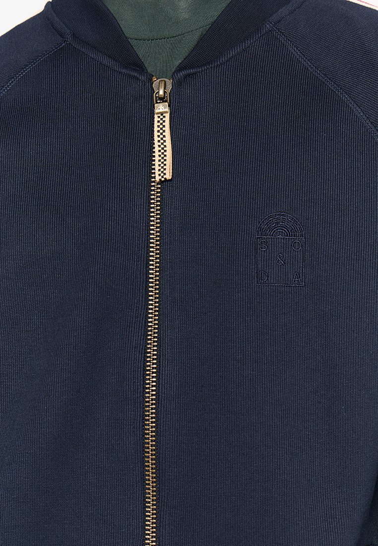 Marineblaue Zip-Jacke mit geripptem Textur, goldenen Reißverschluss und gesticktem Logo. Mit Stehkragen und Kontraststoff innen.
