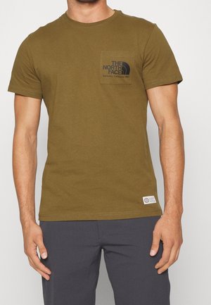 Man draagt een olijfgroen T-shirt met korte mouwen en borstzak met zwart "The North Face"-logo, gecombineerd met donkergrijze broek.
