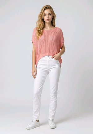 Joven mujer posa con una blusa rosa de punto suelta, vaqueros blancos de talle alto y zapatillas blancas frente a un fondo claro y liso.
