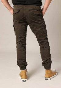 Bruine cargobroek met enkelmanchetten, meerdere zakken en een gladde textuur. Draag met tan kleurige hoge sneakers met witte veters en accenten.