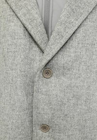 Abrigo de lana gris claro con dos botones marrones y una cremallera plateada visible debajo, mostrando detalles de ropa de invierno en capas.