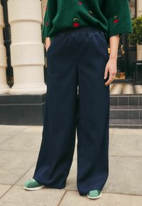 Pantaloni largi de marină, confecționați dintr-un material moale, cu un tiv elastic, asortați cu adidași verzi, cu talpă albă și detalii cu șireturi.