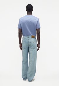 Hombre visto desde atrás con una camisa de manga corta azul clara, vaqueros sueltos de lavado claro y zapatillas blancas sobre un fondo blanco liso.
