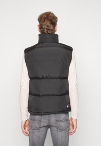 Schwarze Steppweste mit hohem Kragen und horizontaler Steppung. Verfügt über ein Logo-Patch auf der unteren rechten Seite. Getragen über einem hellen Langarmshirt.