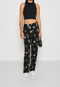 Femme portant un crop top noir sans manches, un pantalon large à fleurs noirs, des baskets blanches, tenant un petit sac à main noir contre un fond uni.