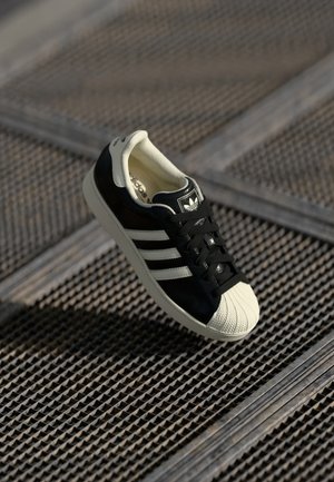 Sort-hvid Adidas-sneaker med skal-tå og tre striber, placeret over en tekstureret metalristflade.