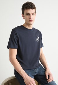 Námorně modré bavlněné tričko s kulatým výstřihem, krátkými rukávy a bílým štítem na levé části hrudi. Hladká textura, volný střih.