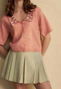 Kobieta ubrana w różowy, zapinany na guziki, krótki kardigan z haftem kwiatowym na kołnierzu oraz jasnobeżową spódnicę plisowaną.
