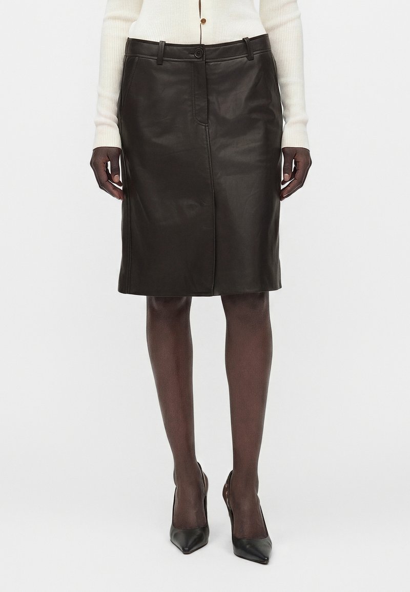Femme portant une jupe en cuir noire longueur genou, des collants noirs, des talons pointus noirs et un haut blanc à manches longues côtelé avec des boutons.