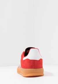 Röda mockaskor med vita läderdetaljer och gummisula. Har diagonala vita ränder och en rundad, vadderad häldesign.