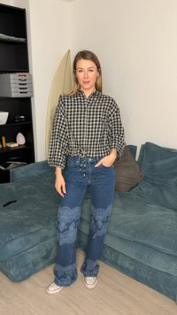 Kobieta stoi w salonie, nosząc czarno-białą kratkowaną bluzkę, niebieskie szerokie spodnie dżinsowe z wzorem oraz białe trampki, za nią znajduje się deska surfingowa i kanapa.