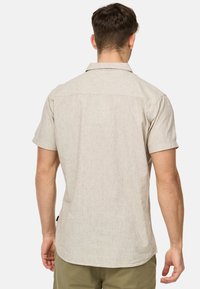 Camisa de manga corta abotonada en tela beige clara con una textura sutil. Presenta un cuello clásico y un dobladillo curvado, mostrando el detalle de la parte posterior.