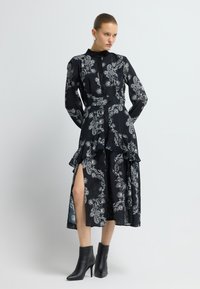 Schwarzes Kleid mit weißen Paisley-Mustern, hohem Kragen, langen Ärmeln, Taillen-Peplum und seitlichen Schlitzen. Kombiniert mit spitzen schwarzen Stiefeletten.