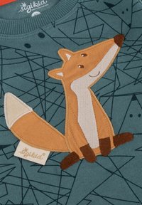 Grüner Stoff-Sweatshirt mit einer bestickten Fuchsfigur, die ein hellbraunes Gesicht, cremefarbene Akzente und einen verspielten Ausdruck hat. Geometrische Muster im Hintergrund.