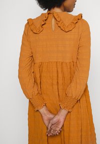 Vestido de manga longa, na cor ferrugem, com um tecido texturizado. Apresenta colarinho com babados e punhos franzidos, acentuando um design em camadas. Detalhe em forma de buraco nas costas.