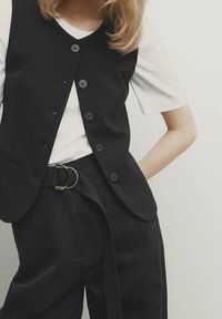 Schwarze Weste mit sechs Knöpfen über einem weißen T-Shirt, kombiniert mit hochtaillierten schwarzen Hosen mit einem Gürtel. Glatter Stoff und taillierte Passform.