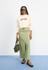 Felpa crema con grafica rosa, gonna midi verde oliva con spacco laterale, scarpe platform leopardate marroni e borsa verde.
