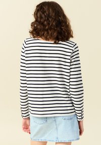 Langermige top in wit en zwarte horizontale strepen, met een ronde hals en knoopaccenten op de mouwen. Spijkerrok zichtbaar daaronder.