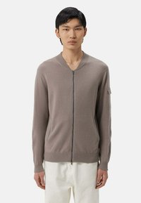 Beige zip-up kofta med ribbad rund halsringning, sidofickor och långa ärmar. Tillverkad av mjukt tyg med en slät yta.