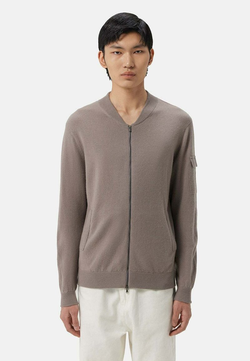 Beige zip-up kofta med ribbad rund halsringning, sidofickor och långa ärmar. Tillverkad av mjukt tyg med en slät yta.