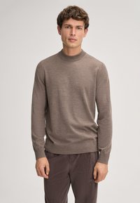 Brązowy sweter dziany z wysokim dekoltem i długimi rękawami, o gładkiej fakturze i dopasowanej sylwetce. Noszony z ciemnymi spodniami z materiału sztruksowego.