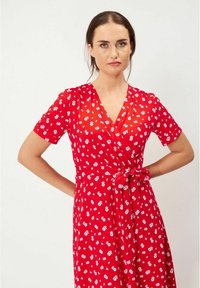 Punainen kukallinen kietaisumekko lyhyillä hihoilla ja vyötäröllä solmimisella. Sen keveässä kankaassa on pieniä valkoisia kukkia. V-pääkohdesign.