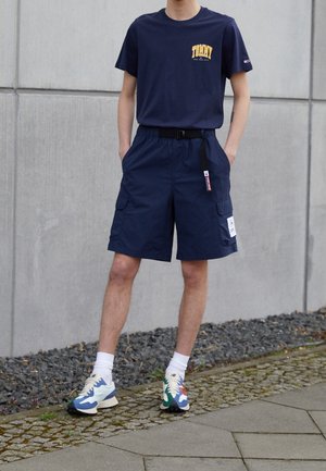 Man draagt een marineblauw grafisch T-shirt, bijpassende cargoshorts met riem, witte sokken en meerkleurige sneakers, staand op de stoep bij een grijze muur.